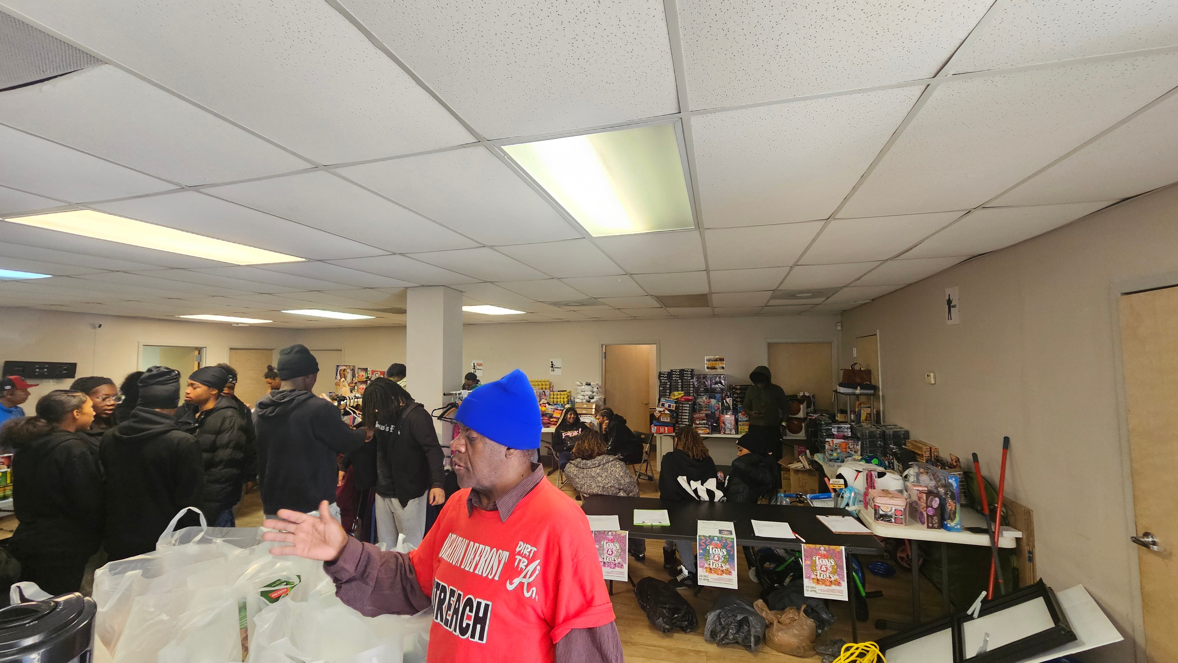People gather in an indoor space with tables and items for sale or display, some wearing hats and jackets.