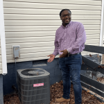 Man standing outdoors next to an air conditioning unit, smiling, wearing a purple shirt and jeans.