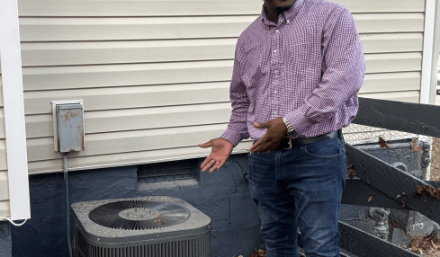 Man standing outdoors next to an air conditioning unit, smiling, wearing a purple shirt and jeans.