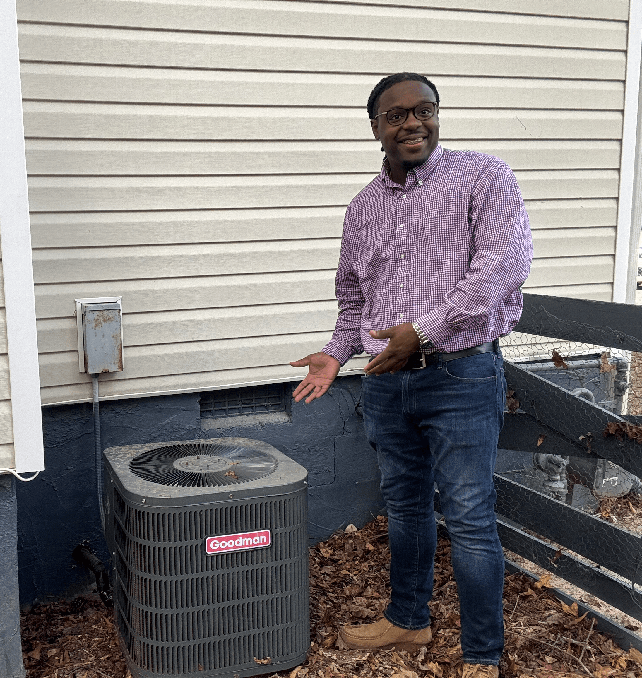 Man standing outdoors next to an air conditioning unit, smiling, wearing a purple shirt and jeans.