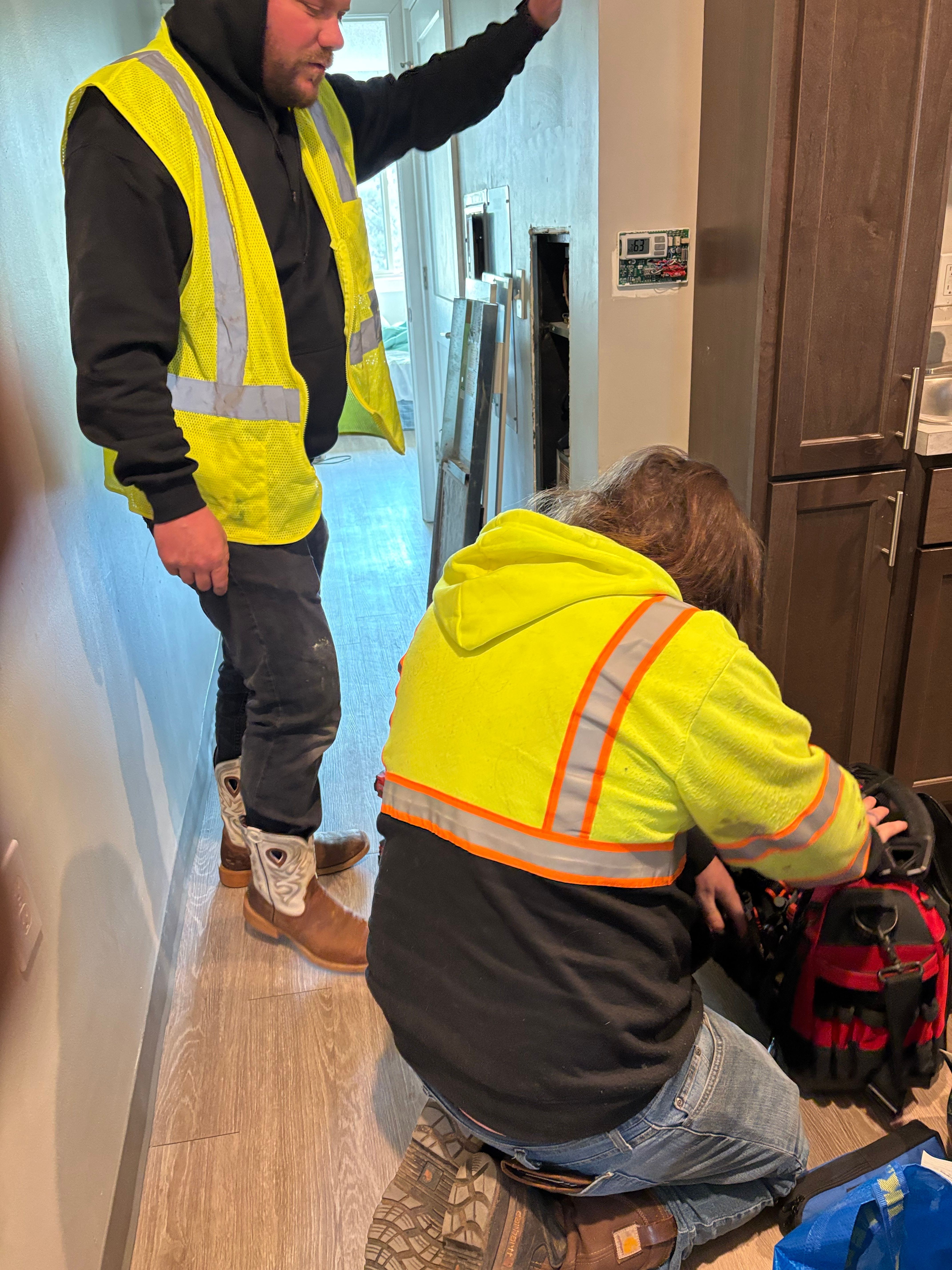 Two people in yellow safety vests and a person in a yellow hoodie with orange reflective stripes are inside a room with wooden cabinets and a window.