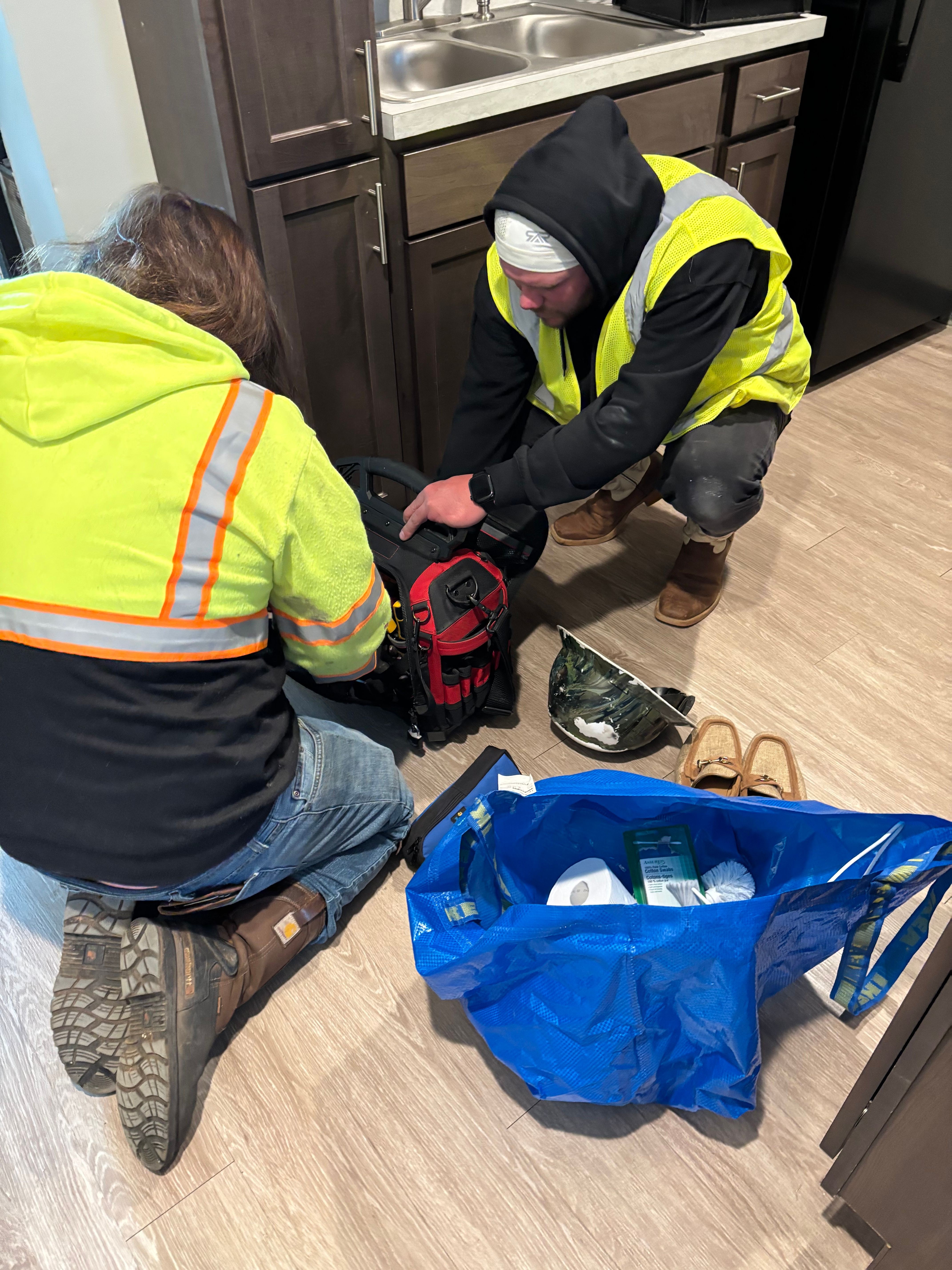 Two workers in safety vests are working on a black bag on the floor, with a blue bag nearby and kitchen cabinets in the background.