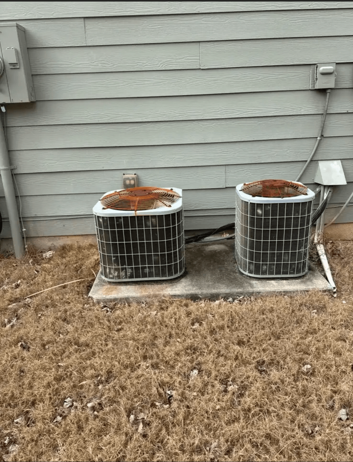 Two outdoor air conditioning units on a concrete pad next to a house with horizontal siding, on a patch of grass.