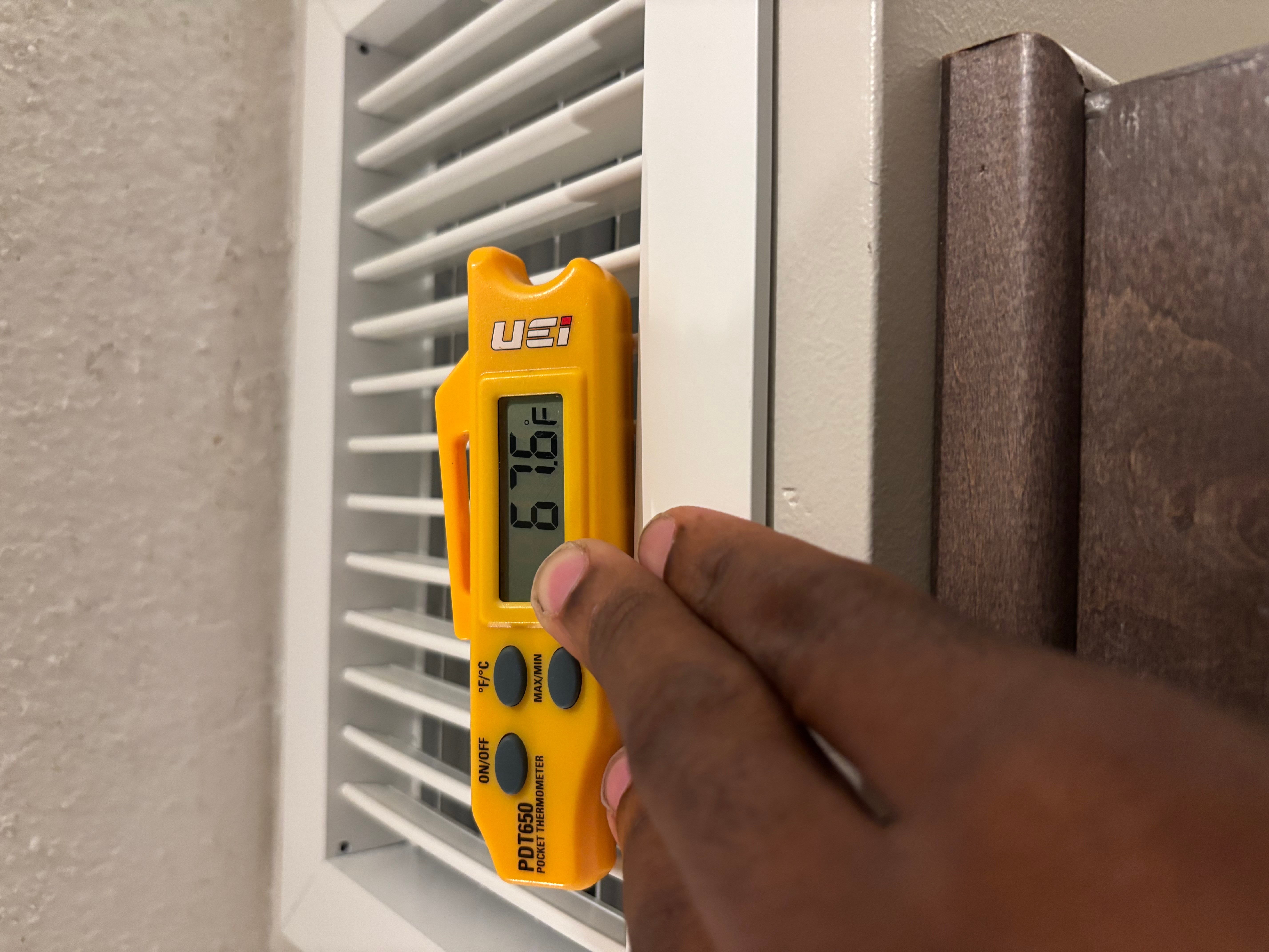 Person holding a yellow infrared thermometer near a white vent, measuring temperature, with a brown surface nearby.