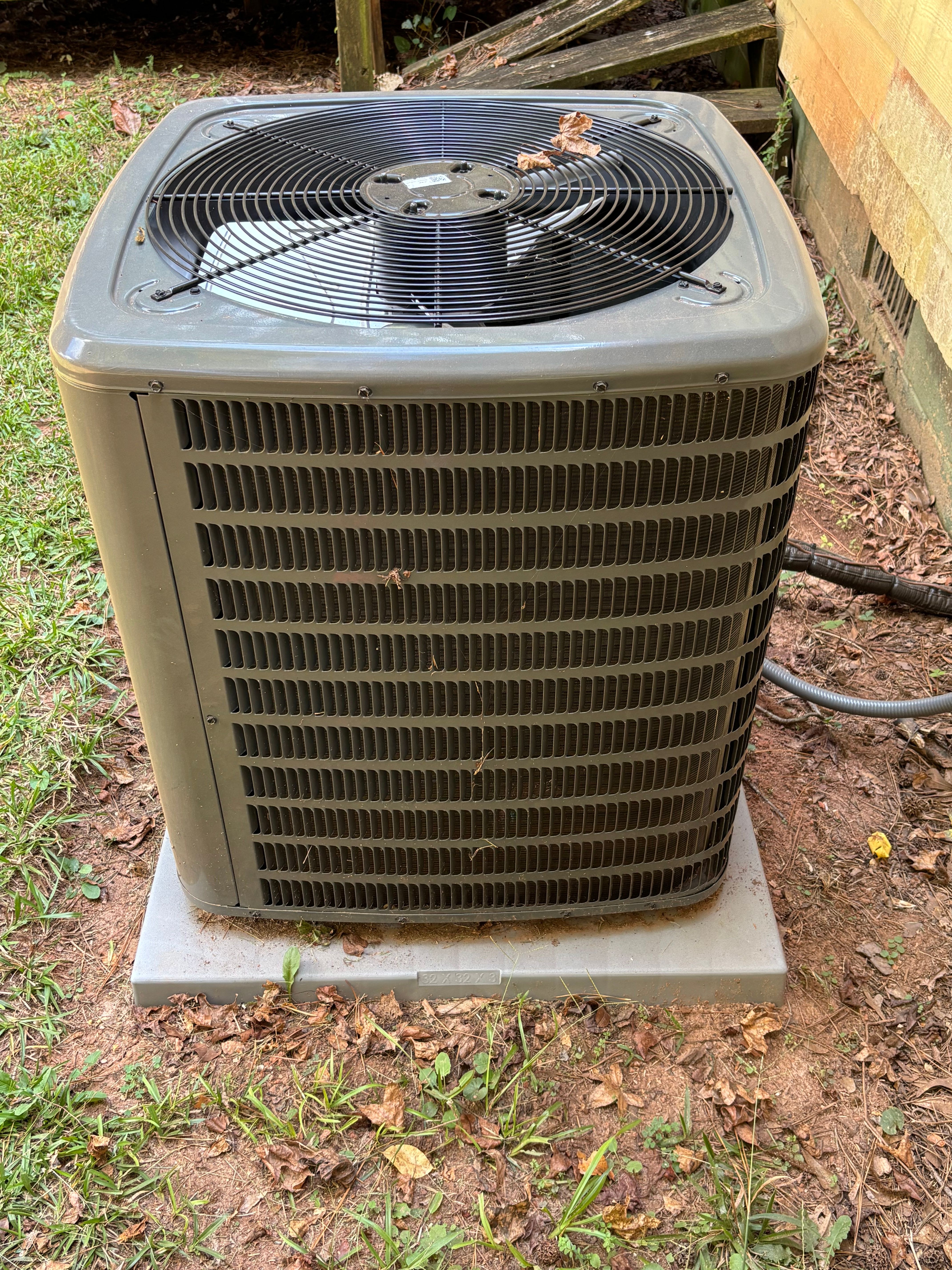 Outdoor air conditioning unit on a concrete pad, surrounded by dirt and plants.