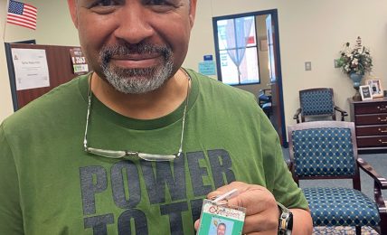 Man with short gray hair and beard smiling, holding an ID badge, in a room with chairs and a clock.