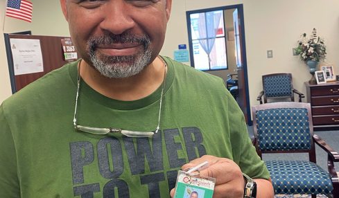Man with short gray hair and beard smiling, holding an ID badge, in a room with chairs and a clock.