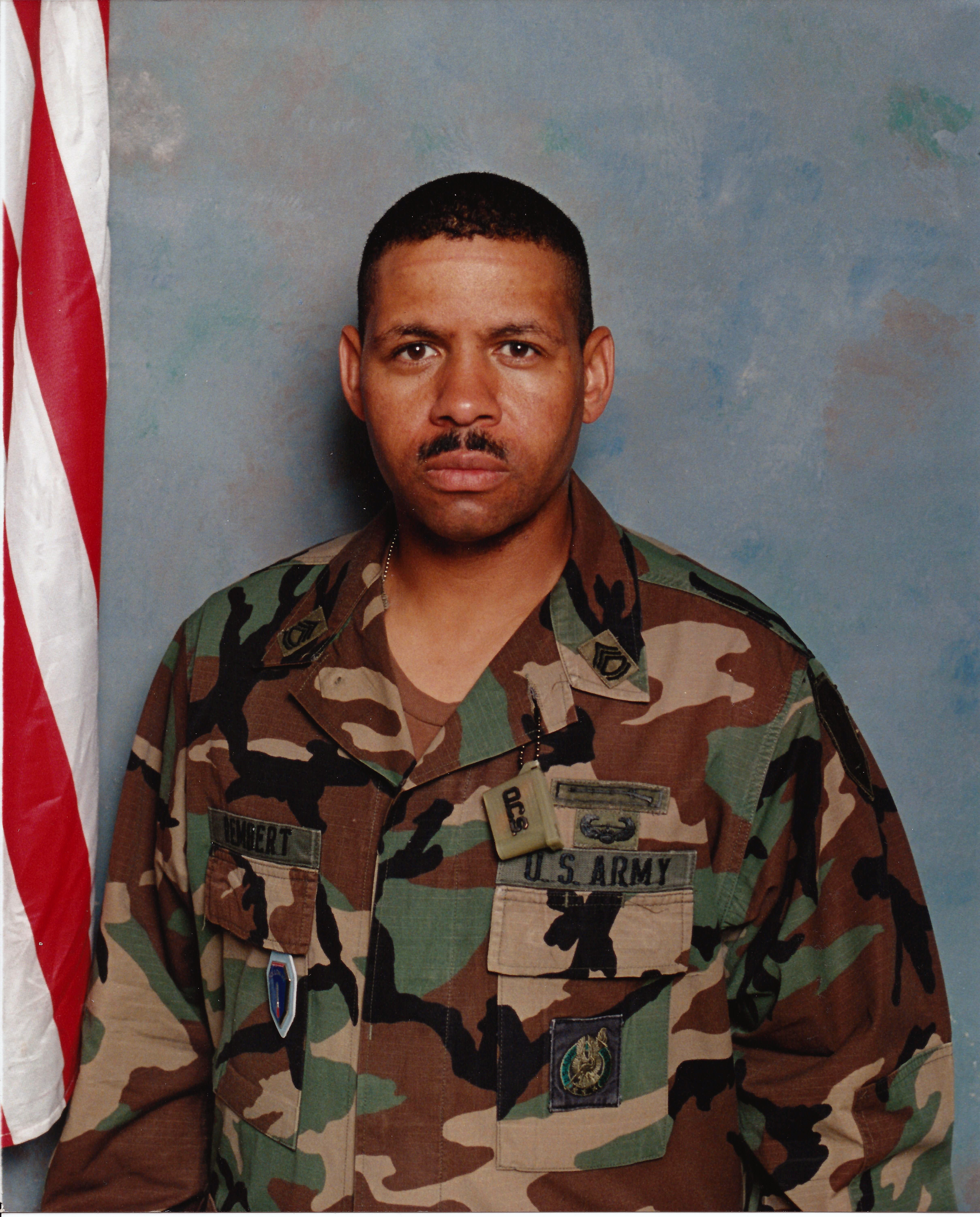 Man in military uniform with camouflage pattern, standing in front of a blue background and American flag, looking at the camera.