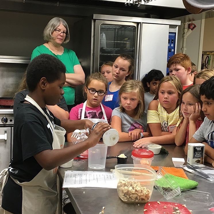 A young person demonstrates a cooking technique to a group of children in a kitchen, with a woman supervising in the background.
