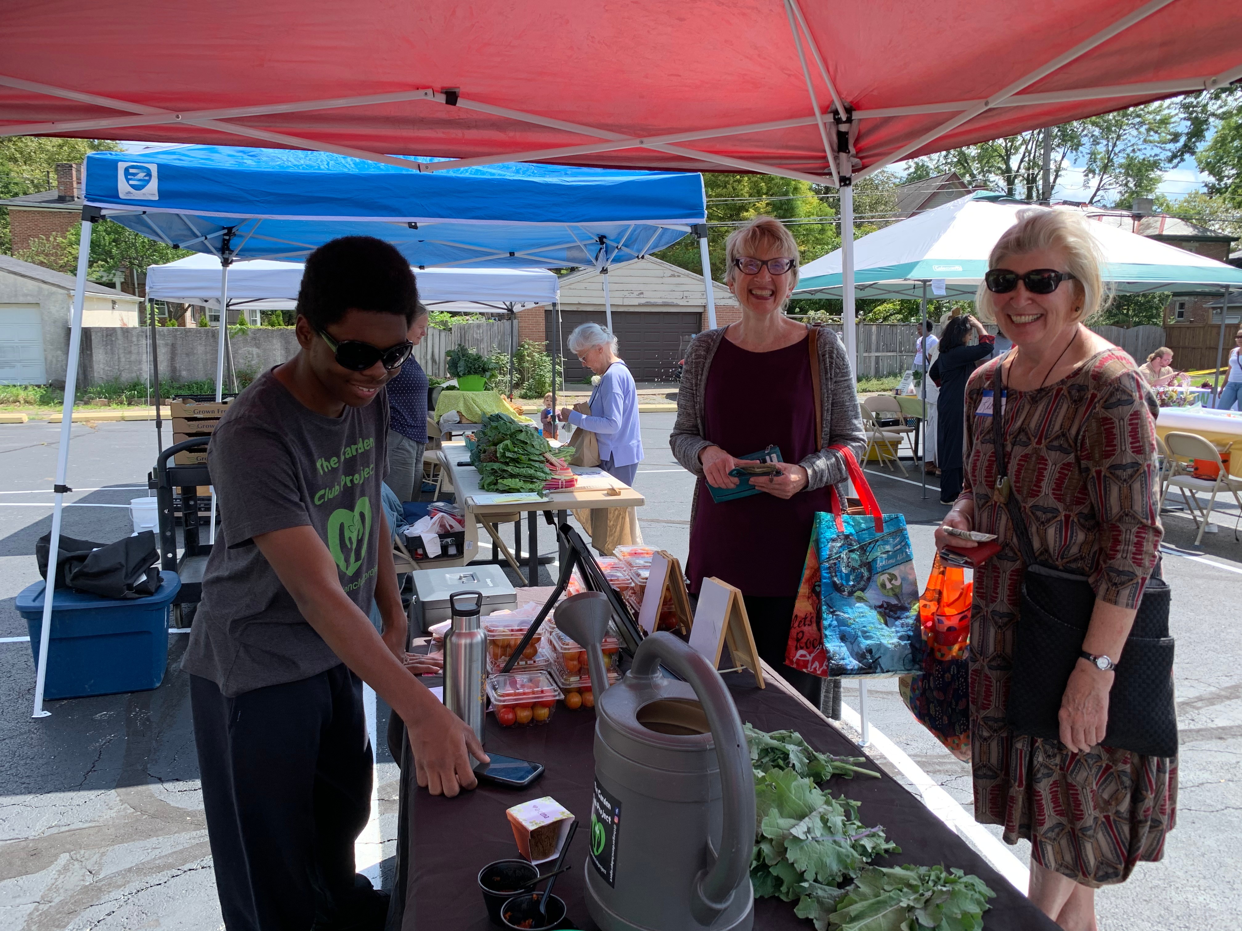 Three people at an outdoor market stall under tents, with tables and plants, smiling and interacting.