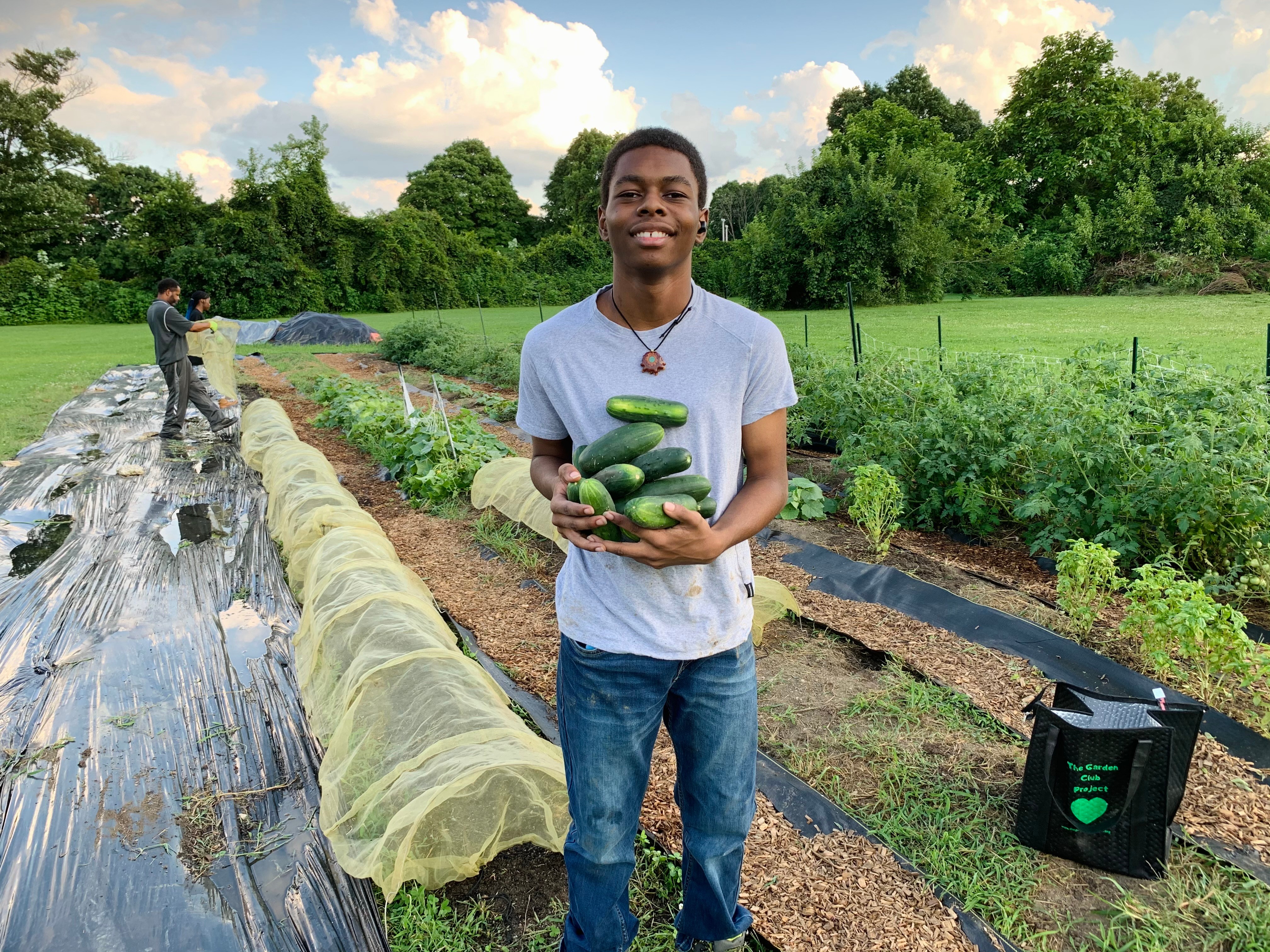 Young man holding cucumbers in a garden with green plants and trees, smiling, sunny day.