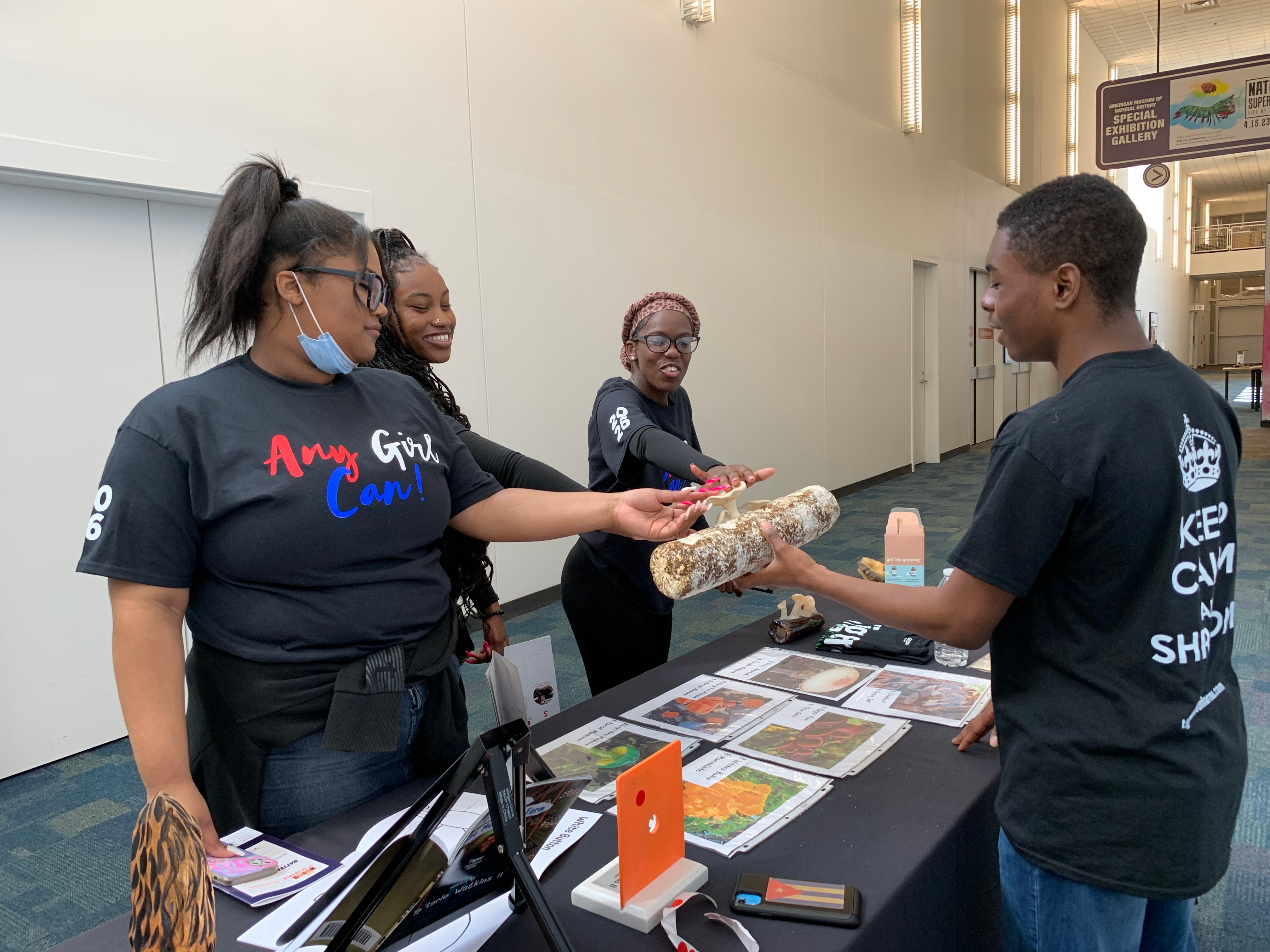 Four women and one man at a table with informational materials, woman handing an item to man, all wearing black shirts.