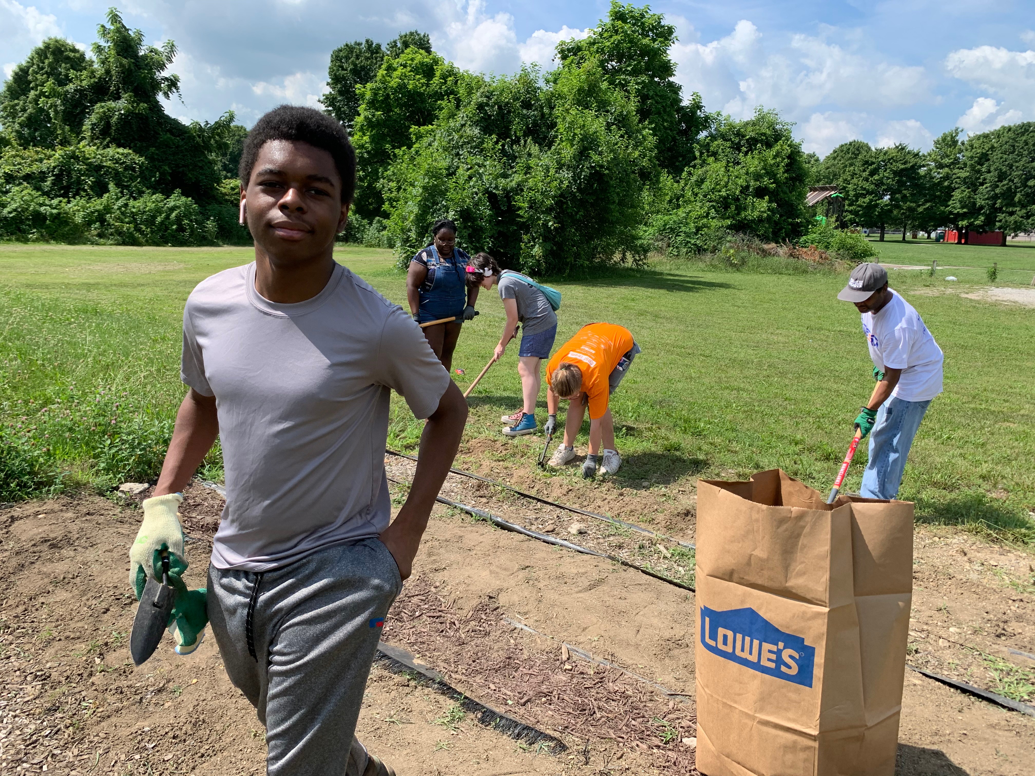 Young man standing outdoors holding gloves, with people working in the background on a grassy field, trees, and a Lowe's shopping bag.