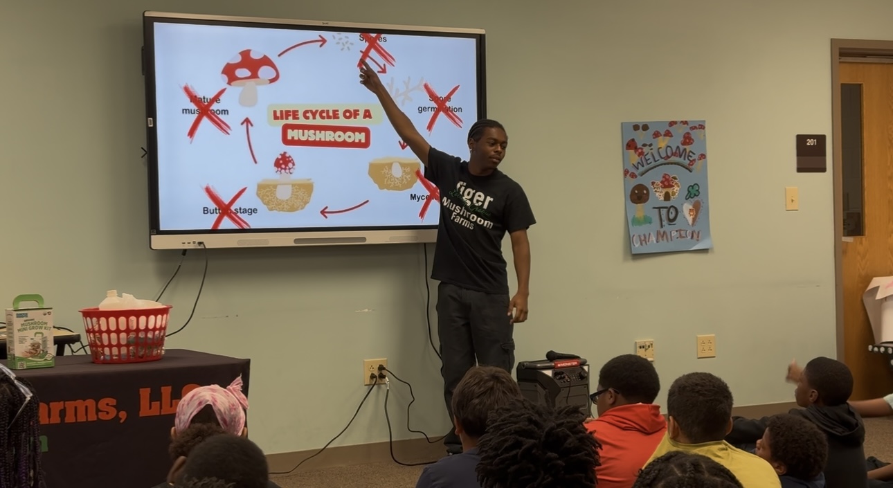 Person standing in front of a screen displaying the life cycle of a mushroom, with children seated and watching.