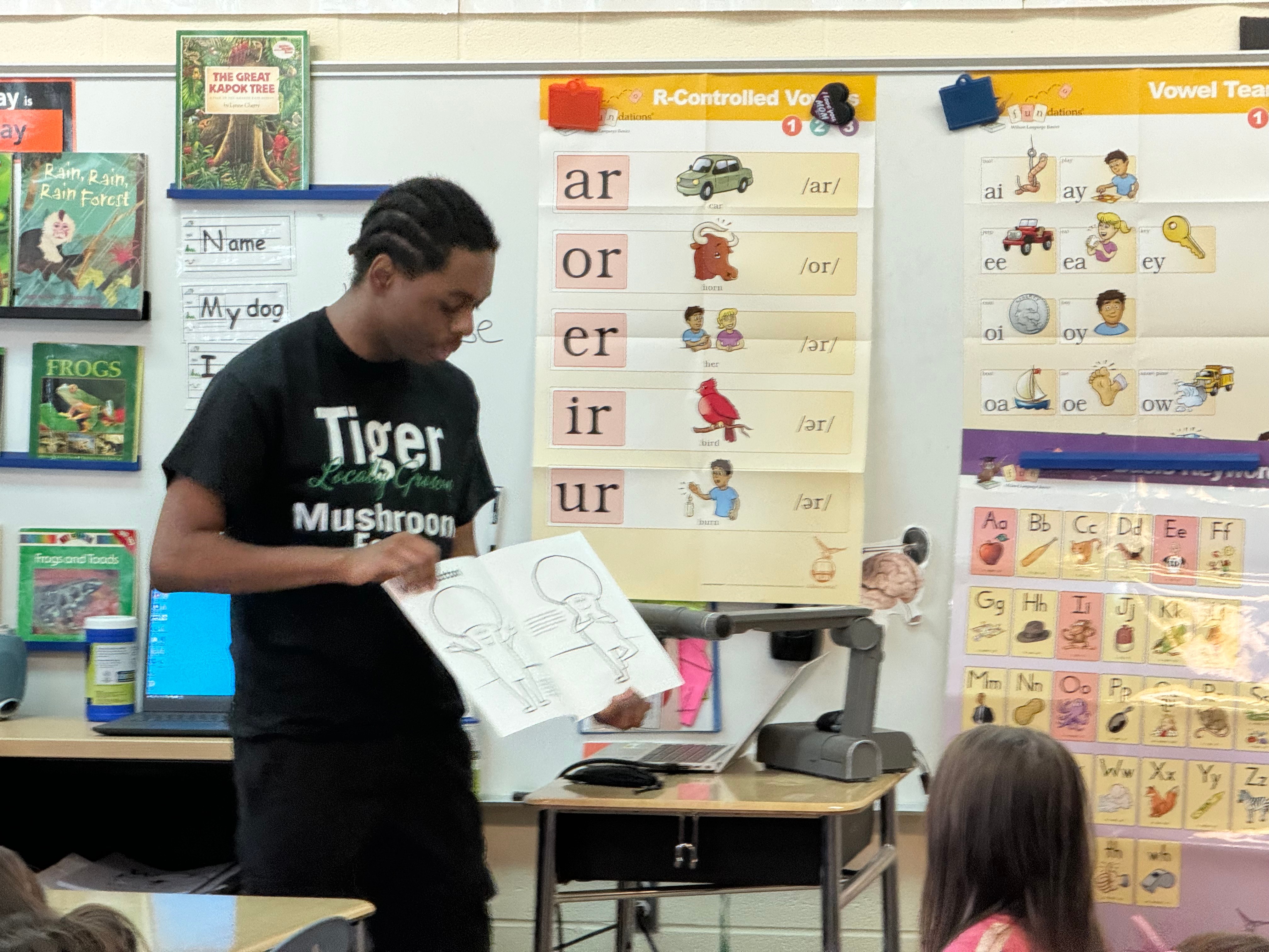 Teacher holding a book in a classroom with educational posters and materials on the wall, students seated nearby.