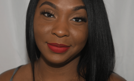 Young woman with straight black hair, red lipstick, and a necklace, smiling at the camera against a plain background.