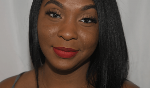 Young woman with straight black hair, red lipstick, and a necklace, smiling at the camera against a plain background.