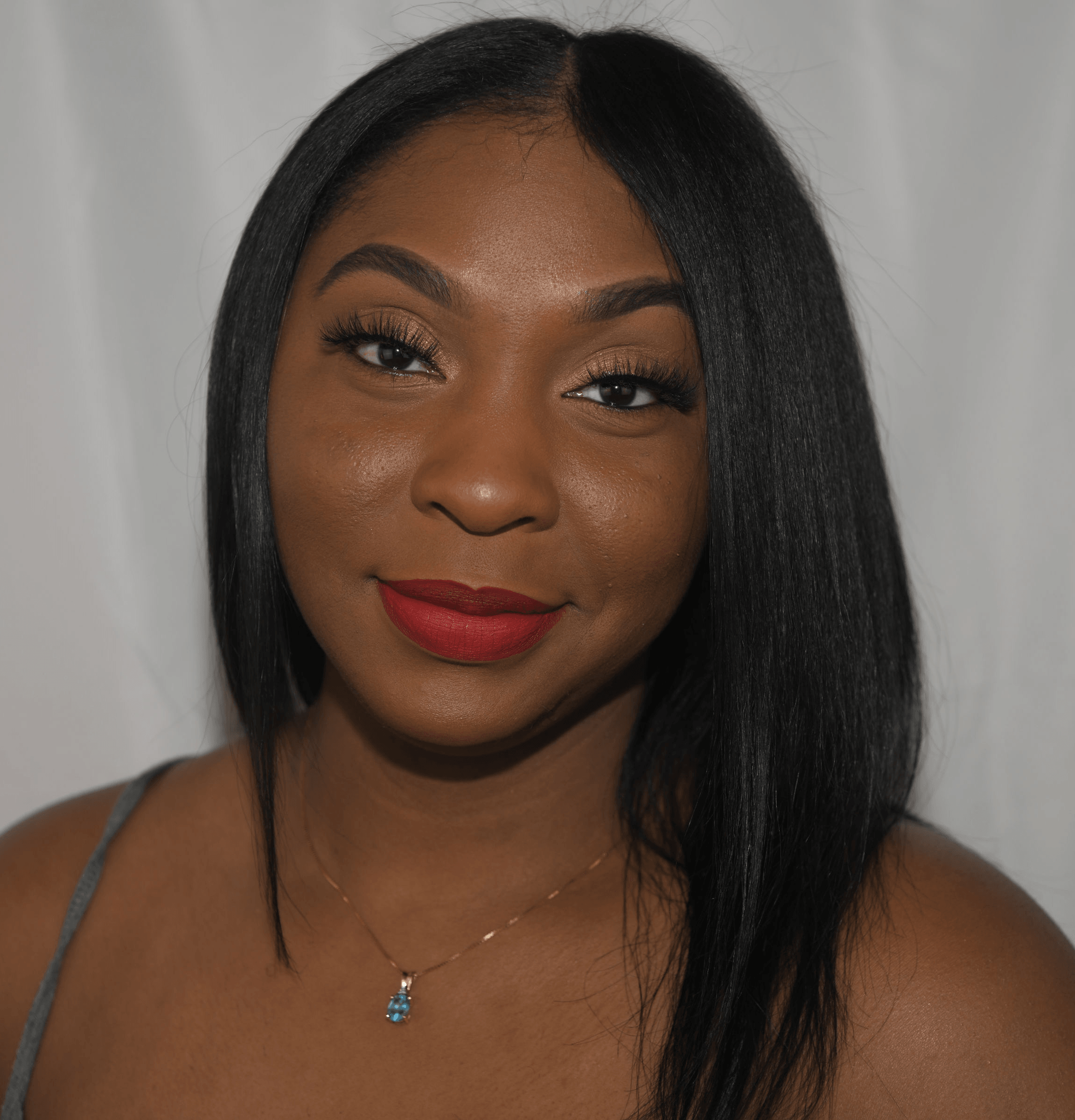 Young woman with straight black hair, red lipstick, and a necklace, smiling at the camera against a plain background.