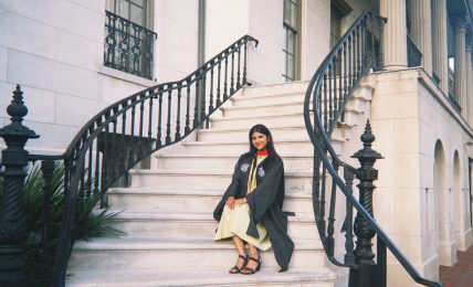Woman in a black coat and yellow dress sitting on white stone stairs outside a building with black railings.