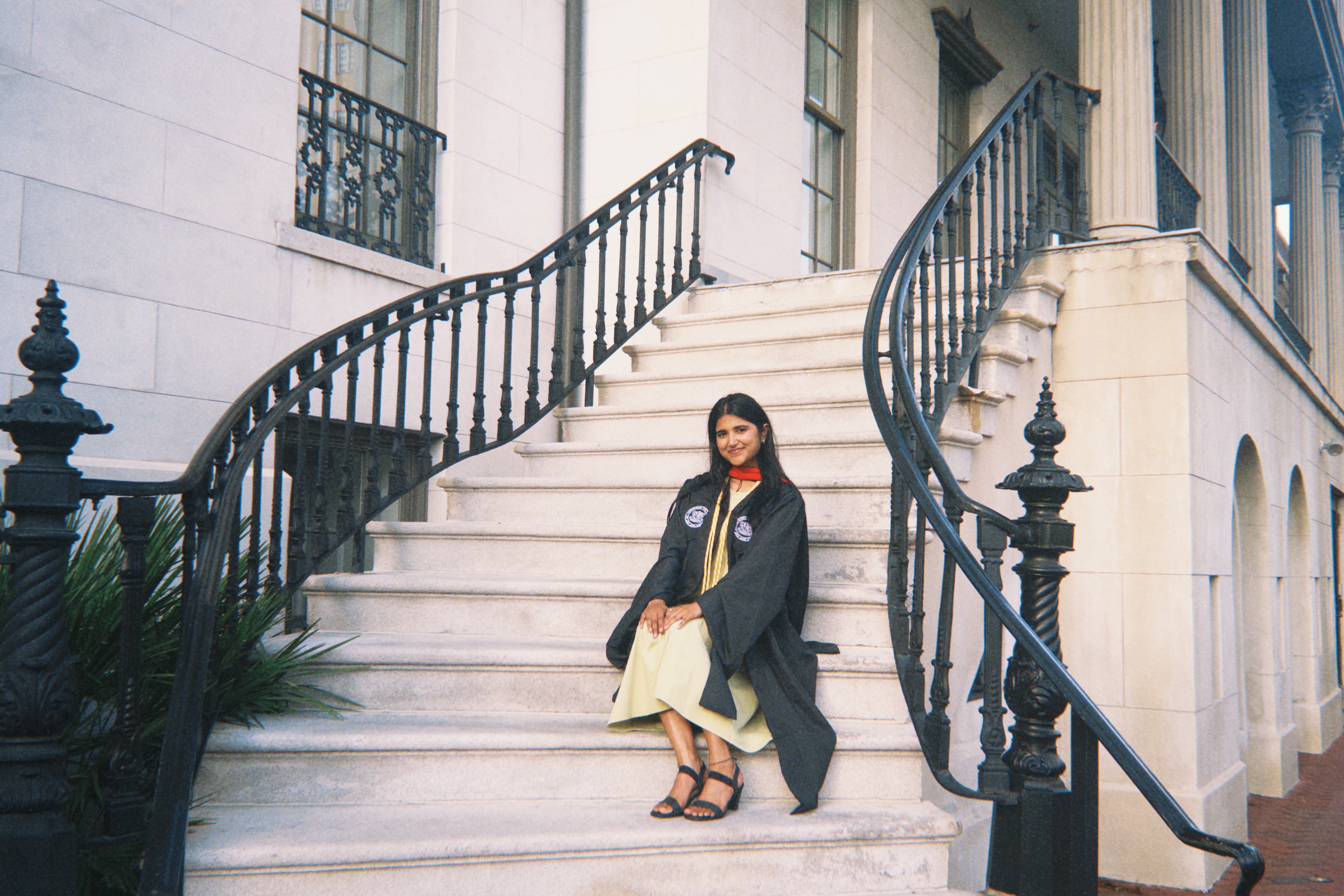 Woman in a black coat and yellow dress sitting on white stone stairs outside a building with black railings.