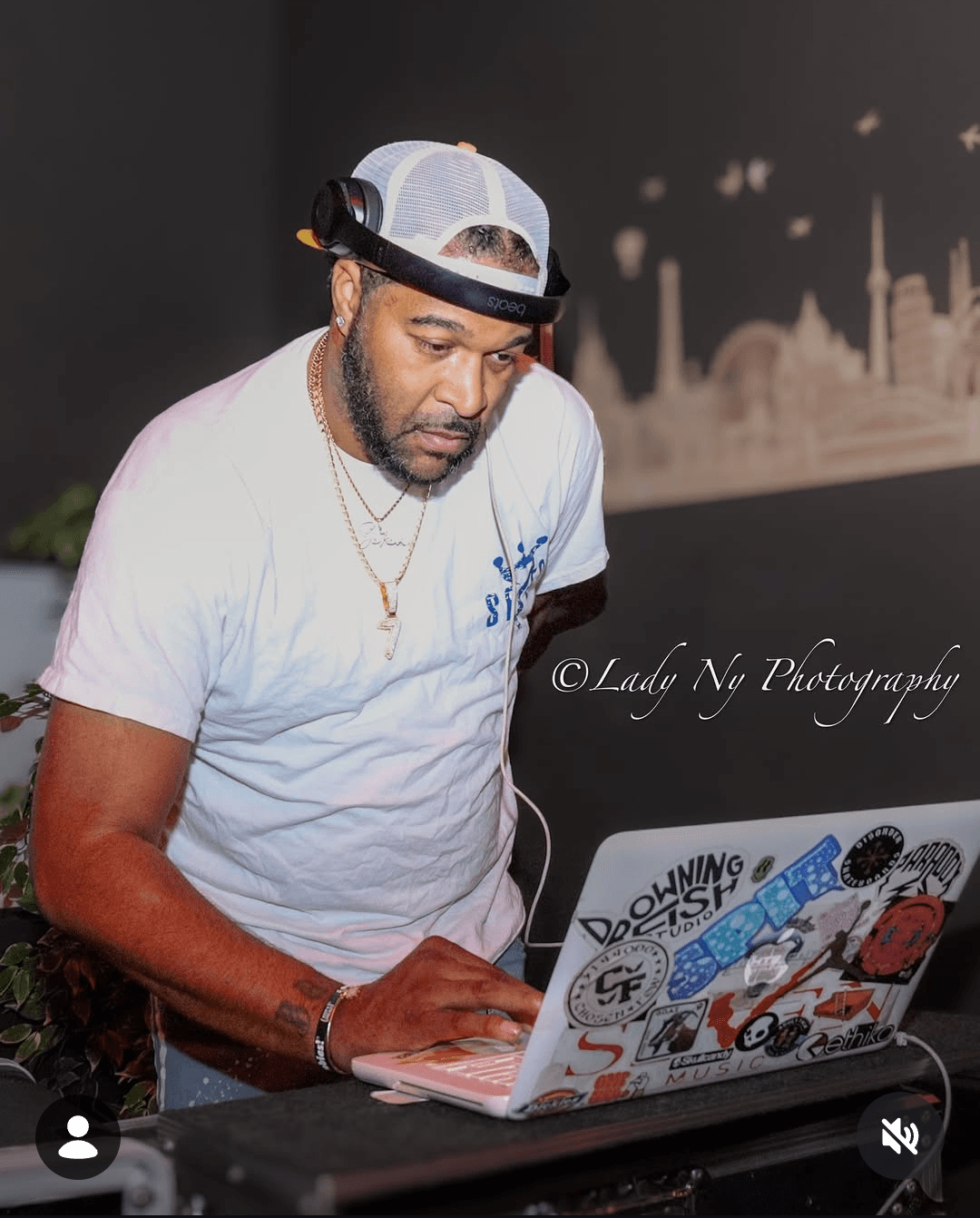 Man wearing a white cap and t-shirt, working on a laptop decorated with stickers, in front of a cityscape background.