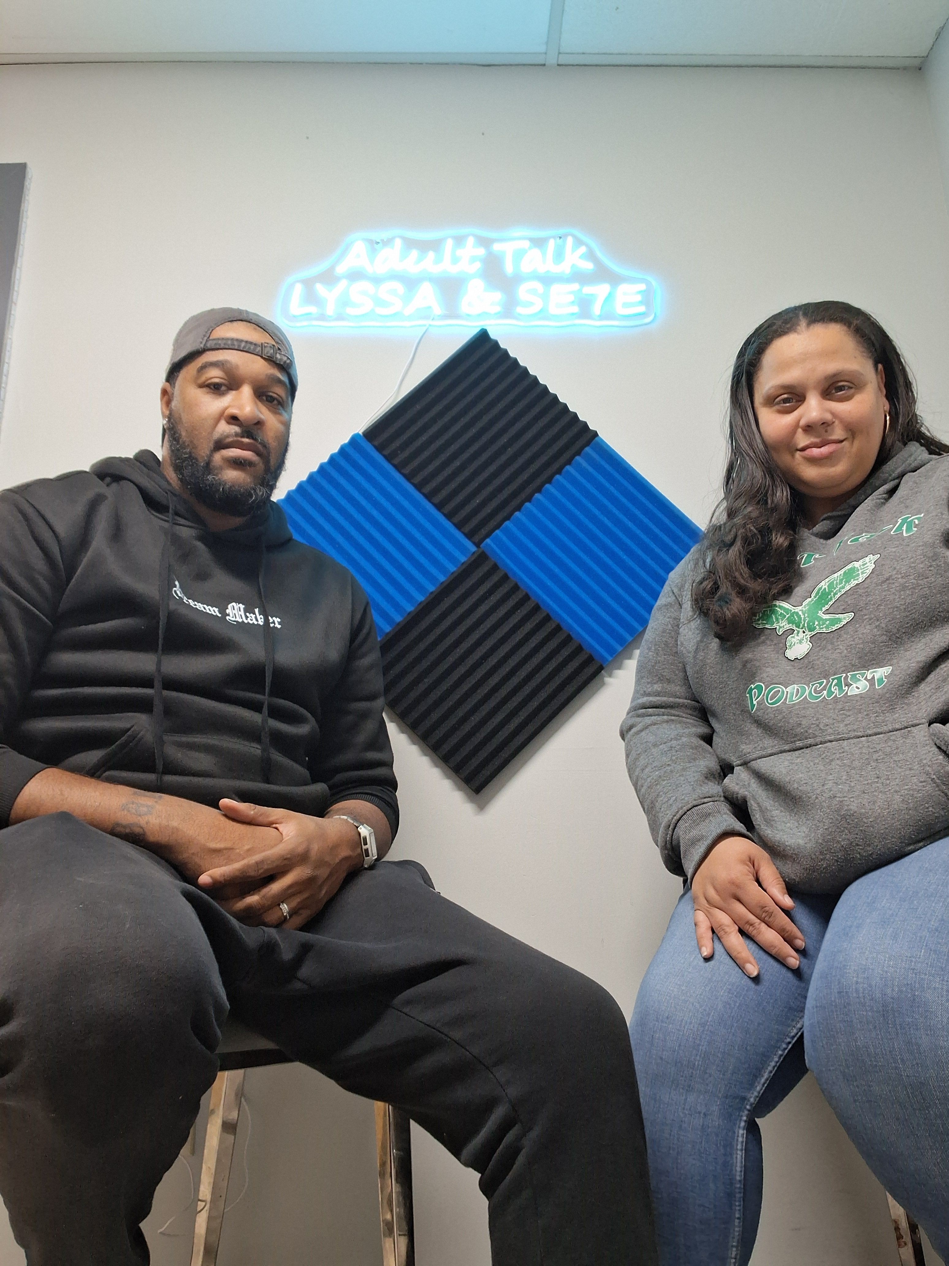 Two people sitting in a room with soundproof panels and a neon sign on the wall, one wearing a black hoodie, the other a gray sweatshirt.