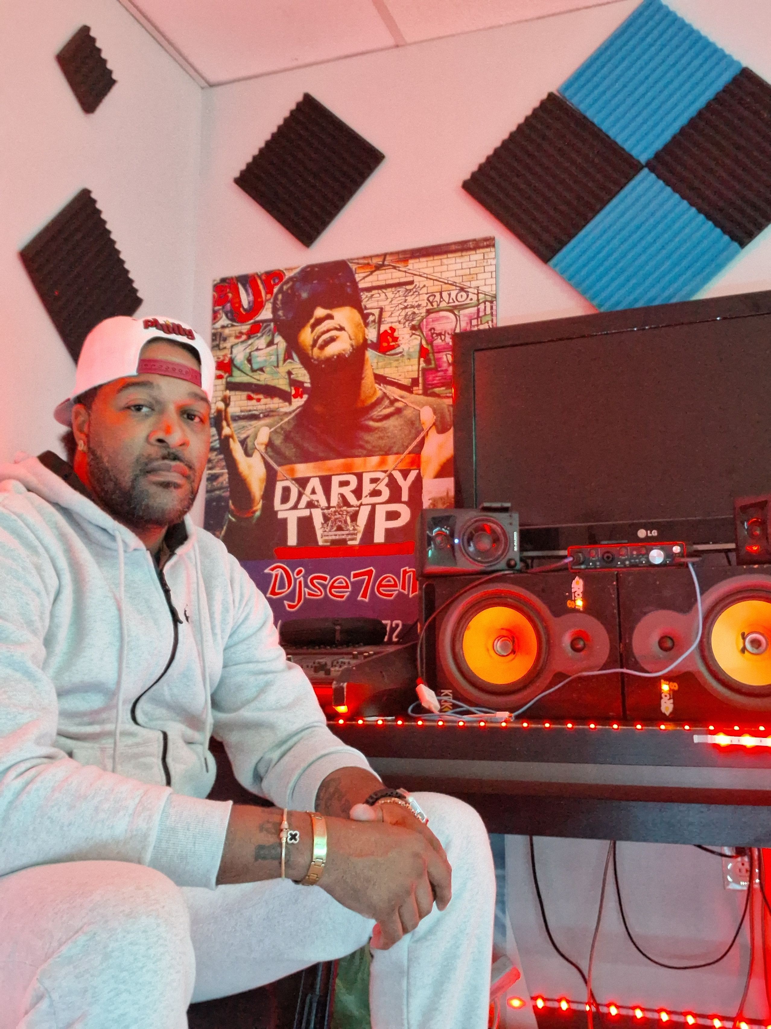 Person in a white hoodie and cap sitting in a music studio with DJ equipment and posters on the wall.