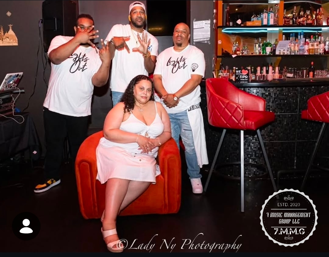 Four people, three men and one woman, pose in a bar with shelves of bottles in the background. The woman sits on a red chair.