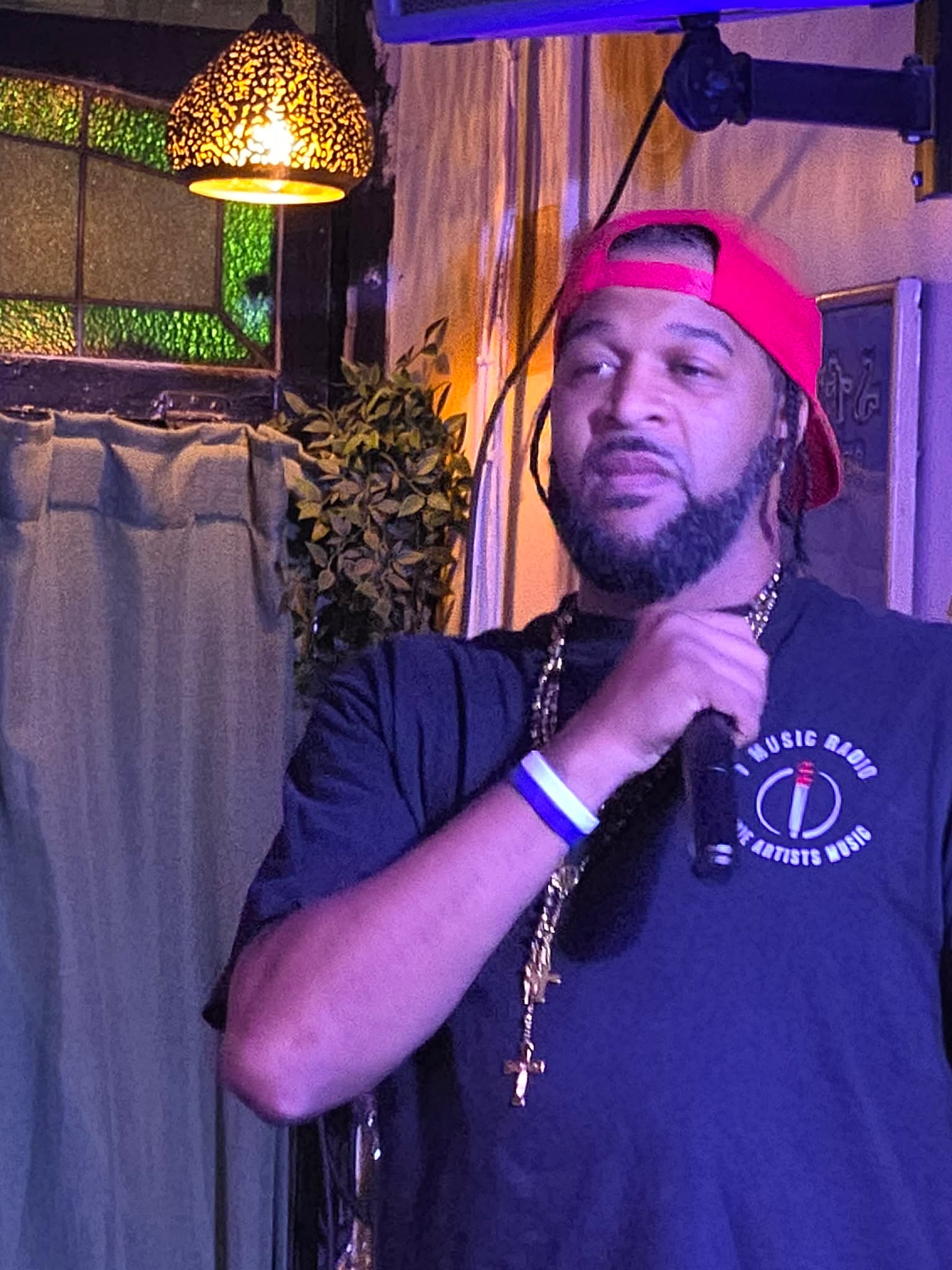 Man with a beard wearing a red cap backwards, black t-shirt, and jewelry, holding a microphone indoors.