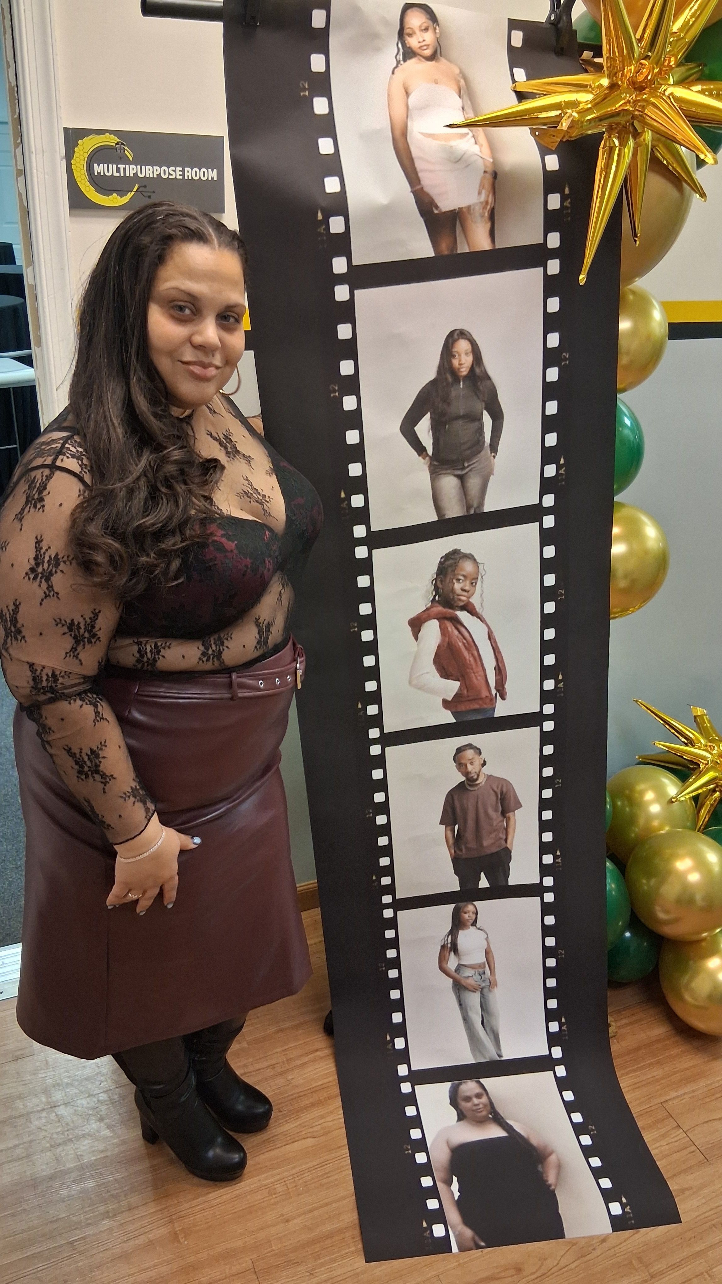 A woman with long dark hair wearing a patterned blouse and brown skirt stands indoors near a photo strip and holiday decorations.