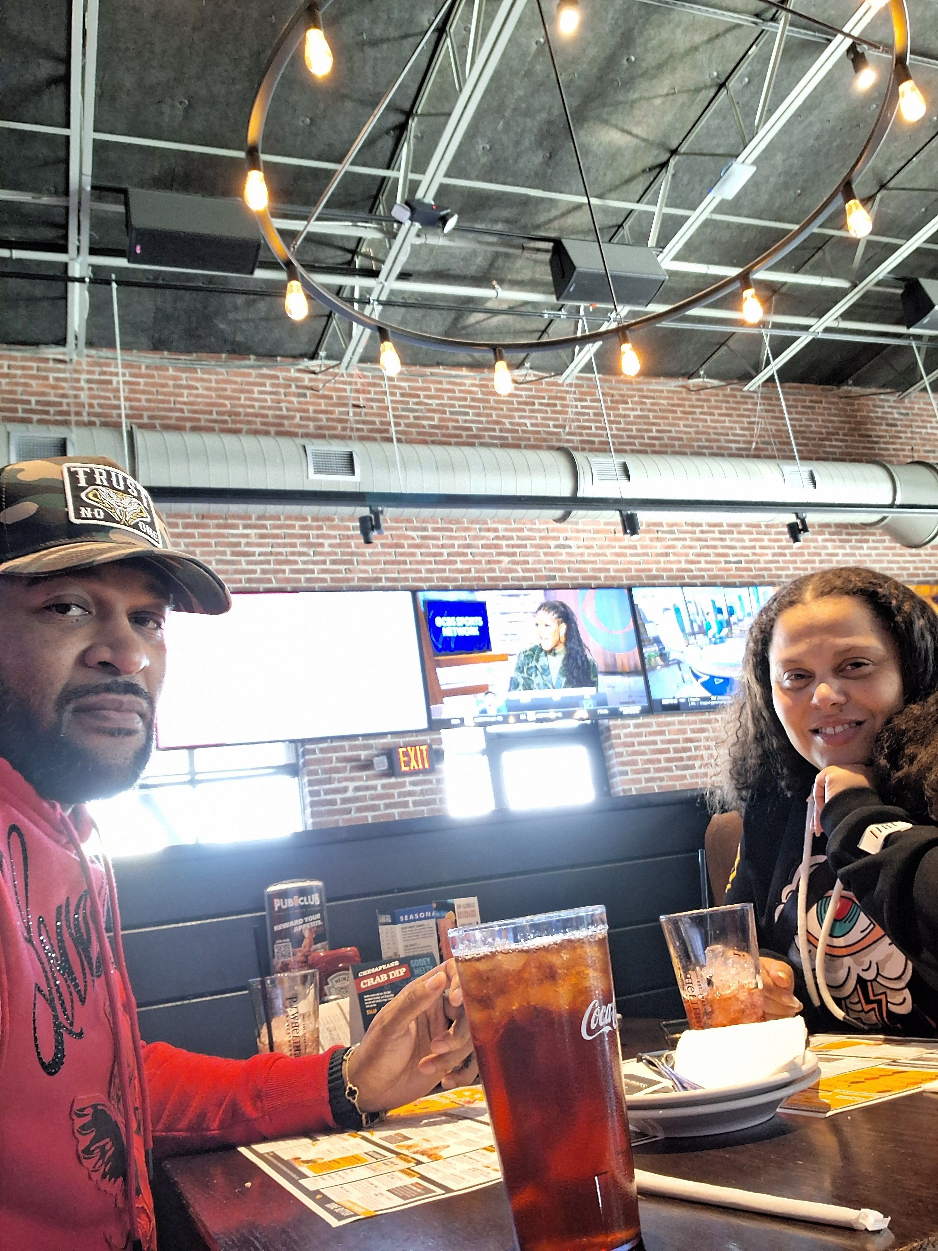 Two people sitting at a bar counter with drinks, in a modern, well-lit setting with large windows and ceiling lights.