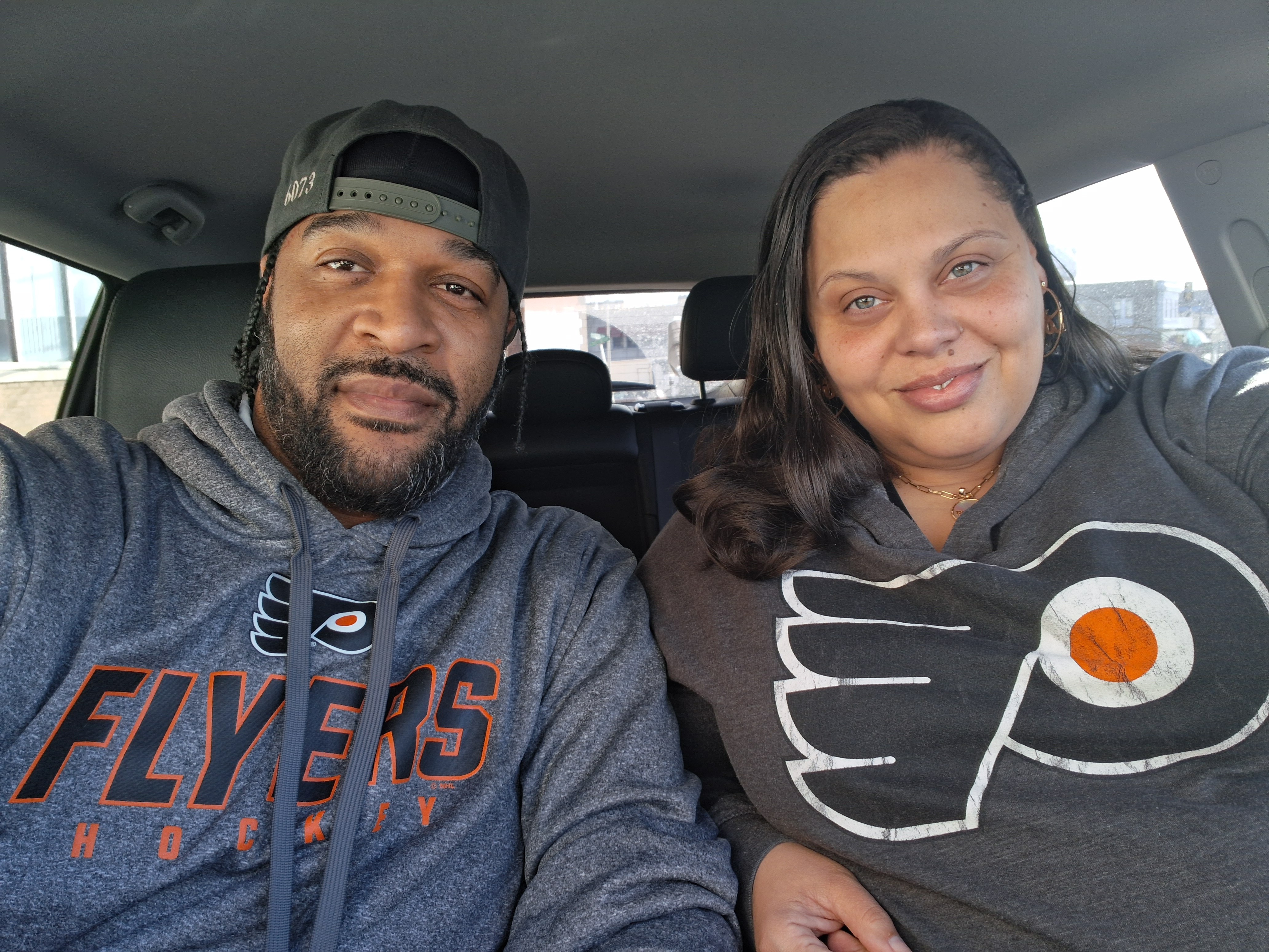 Two people sitting in a car, smiling at the camera, with a city view outside the window.