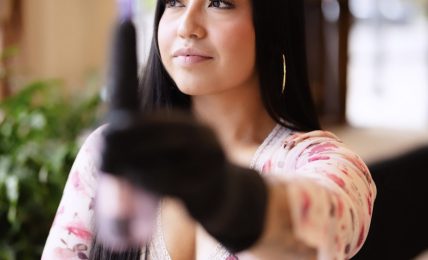 Young woman with long dark hair wearing a floral top, extending her arm towards the camera, in an outdoor setting.