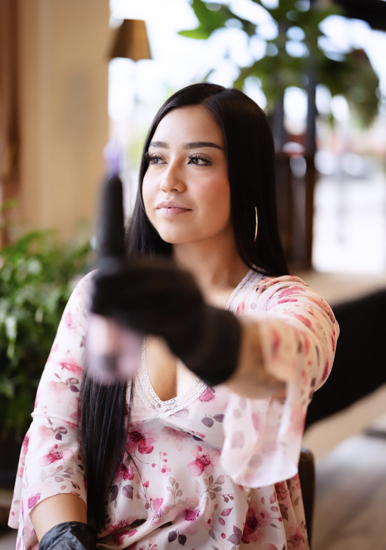 Young woman with long dark hair wearing a floral top, extending her arm towards the camera, in an outdoor setting.