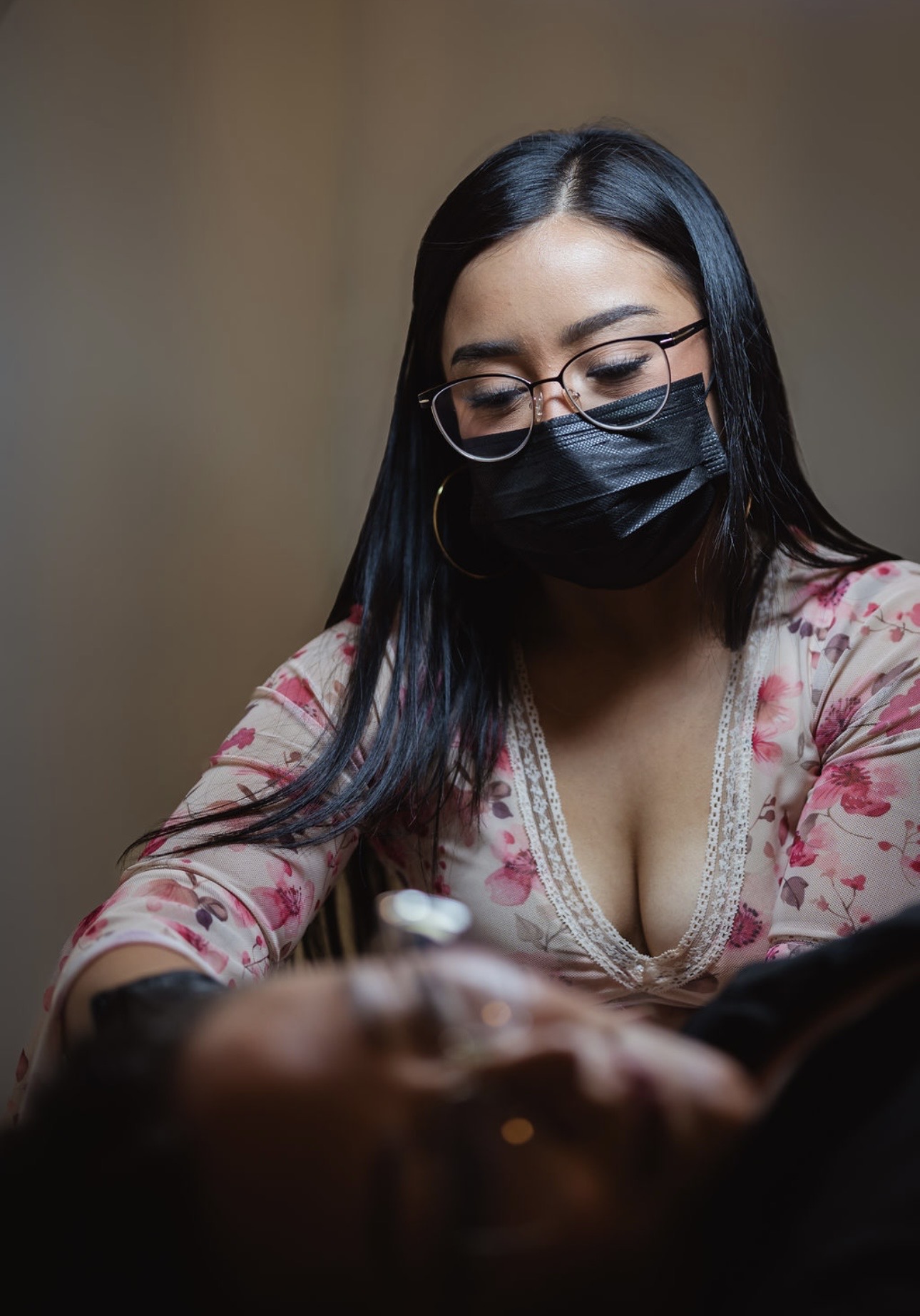 Woman with glasses and black face mask looking down, wearing a floral top, indoors.