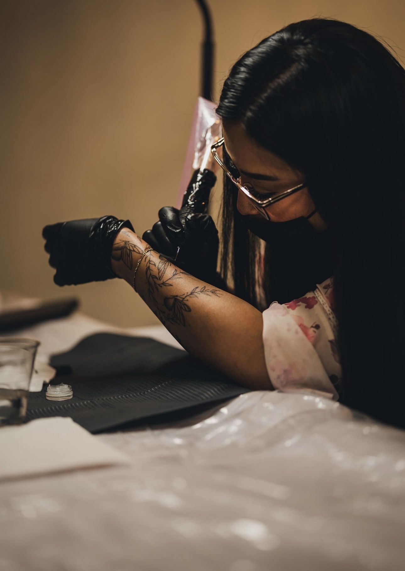 Person with long dark hair and glasses tattooing skin with a tattoo machine, wearing gloves, in a tattoo studio.