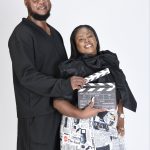 A man and woman smiling, holding a film clapperboard, standing against a plain white background.