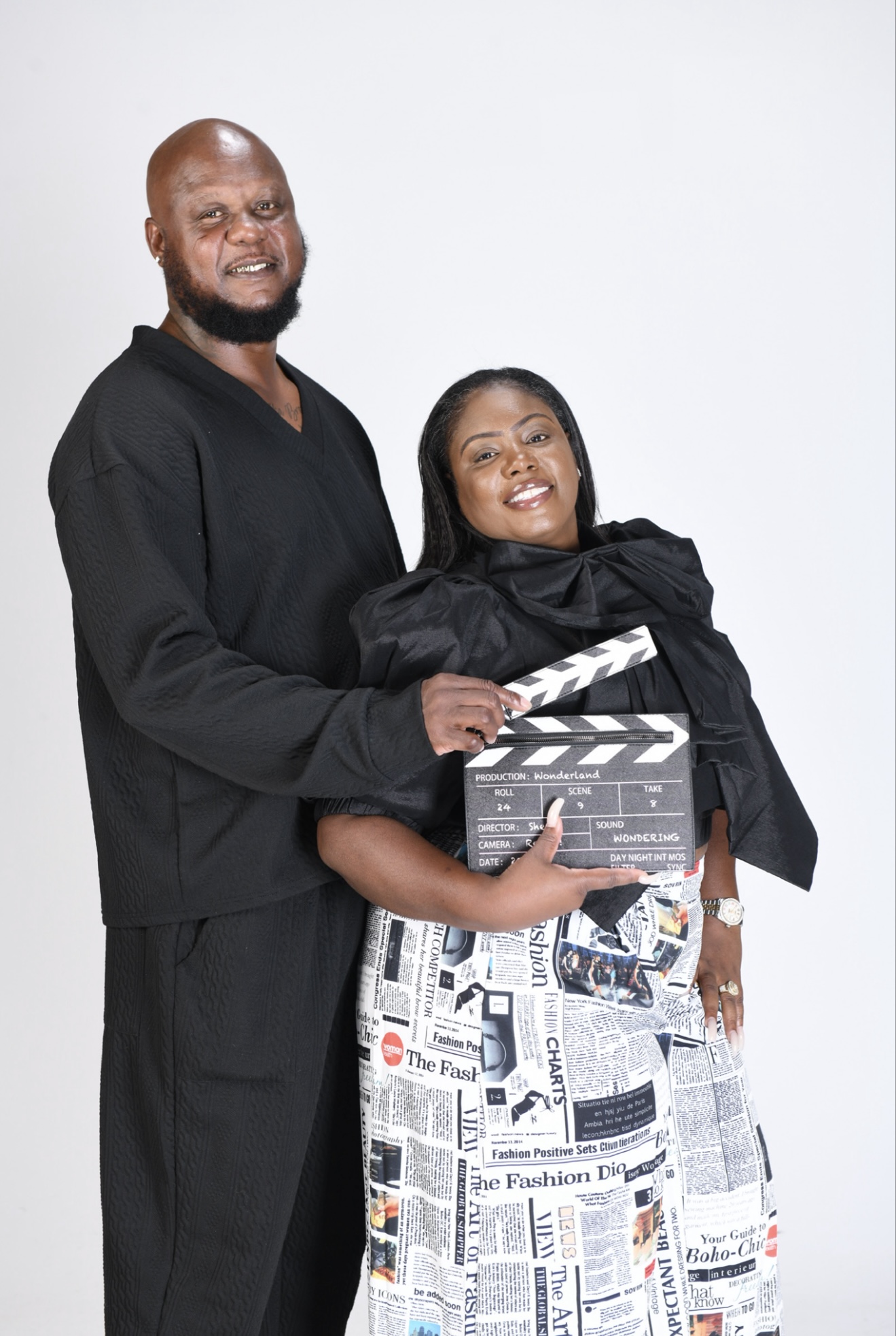 A man and woman smiling, holding a film clapperboard, standing against a plain white background.