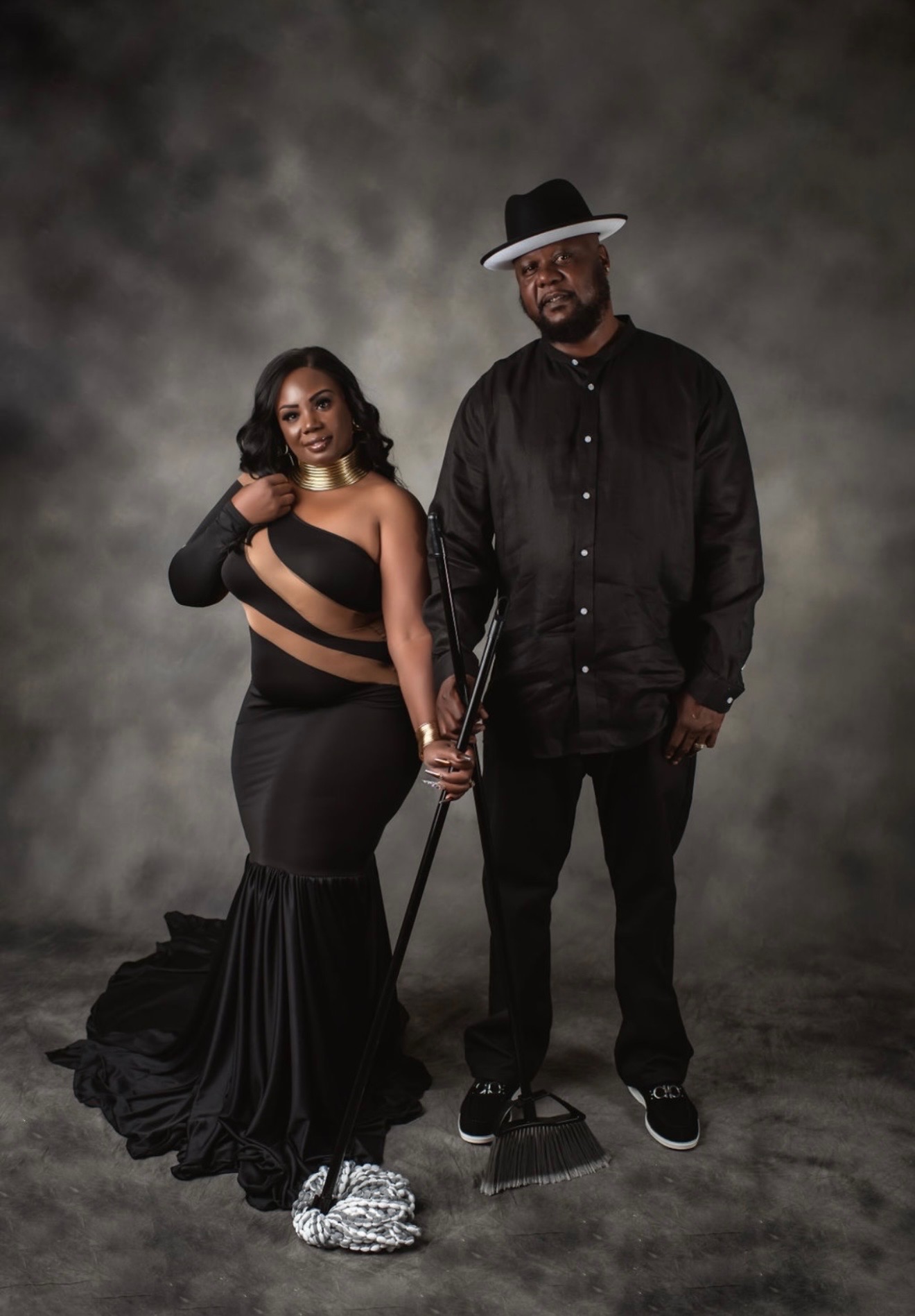 A woman in a black dress with a slit and a man in a black outfit and hat stand together holding a cane, smiling.