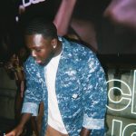 Young man with short hair wearing a blue patterned jacket and white shirt, operating DJ equipment at an event.