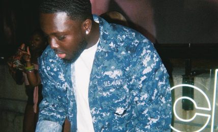 Young man with short hair wearing a blue patterned jacket and white shirt, operating DJ equipment at an event.
