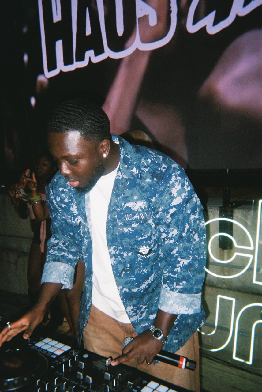 Young man with short hair wearing a blue patterned jacket and white shirt, operating DJ equipment at an event.