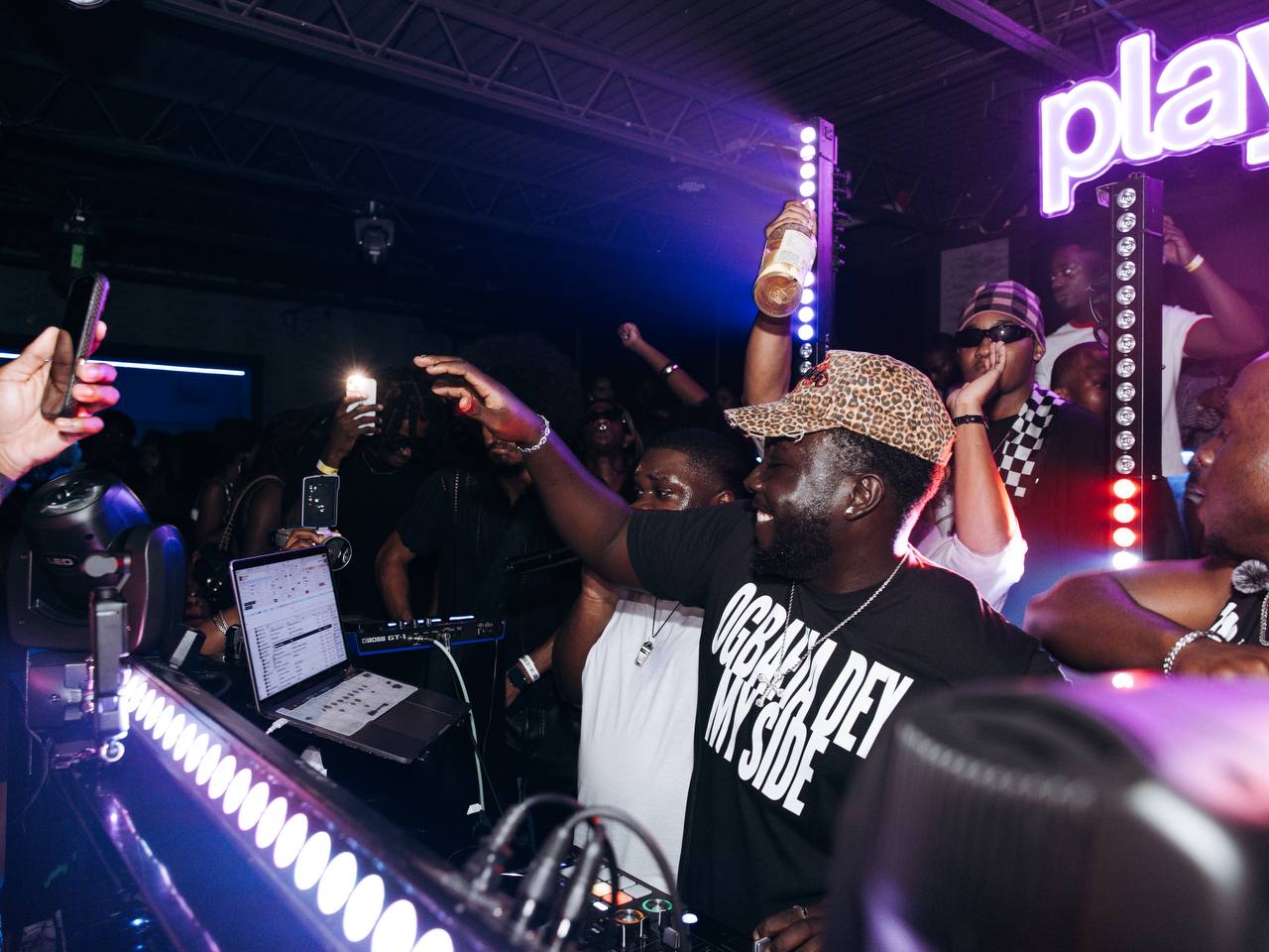 People dancing and taking photos at a nightclub with purple and pink lighting, DJ equipment, and a large illuminated sign reading 'play'.