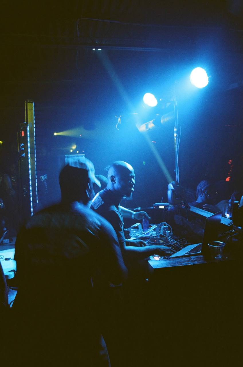 People at a dimly lit nightclub with blue and yellow stage lights, some facing a DJ or performer, dark background.