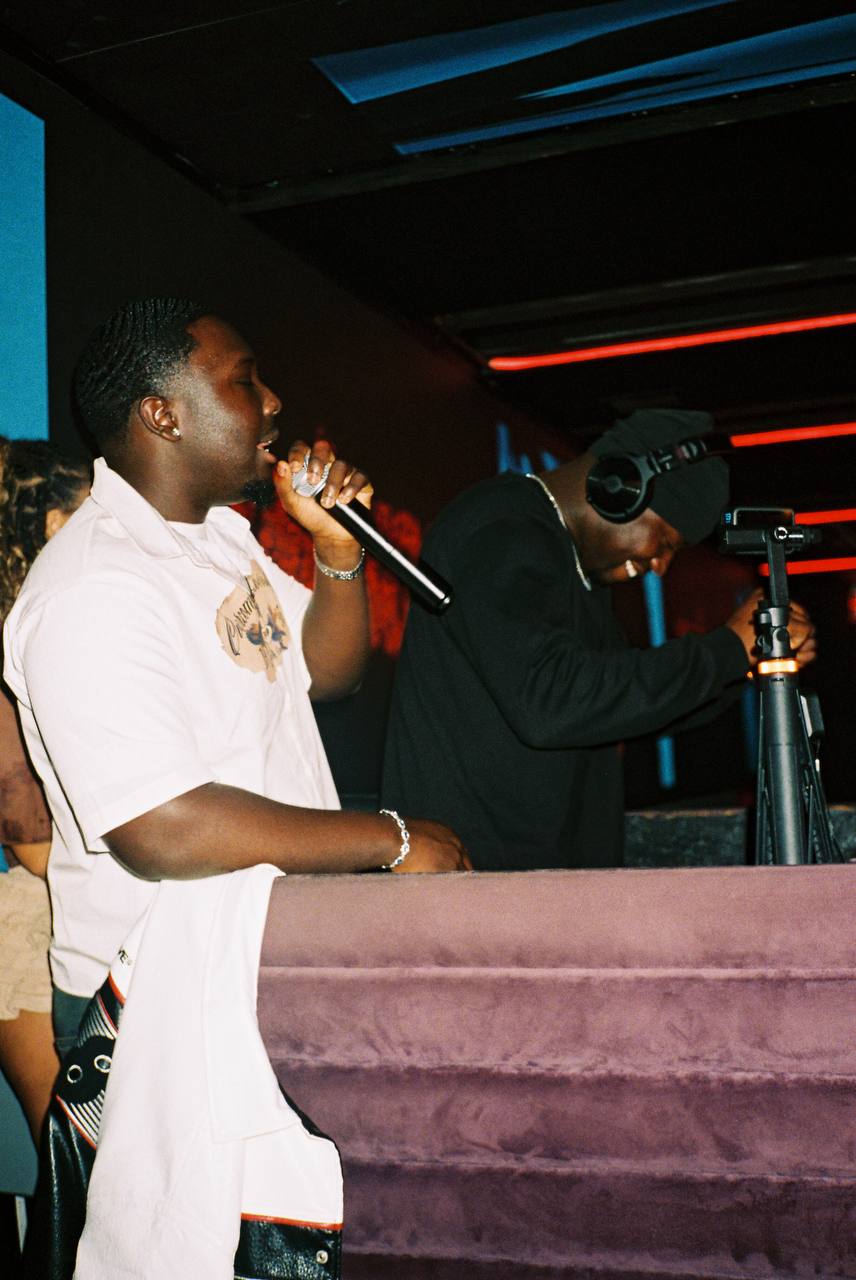 Two men stand at a microphone on a stage with colorful lighting, one holding a microphone and the other adjusting equipment.