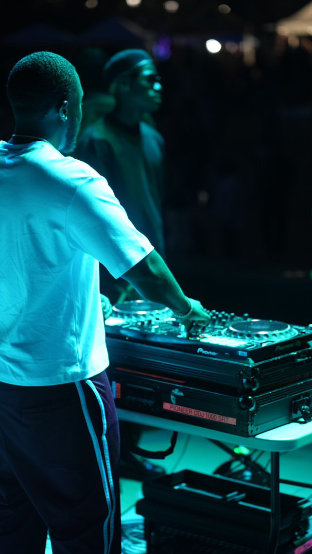 Person DJing with turntable and mixer, illuminated by blue light, with a woman in the background at a dark event.