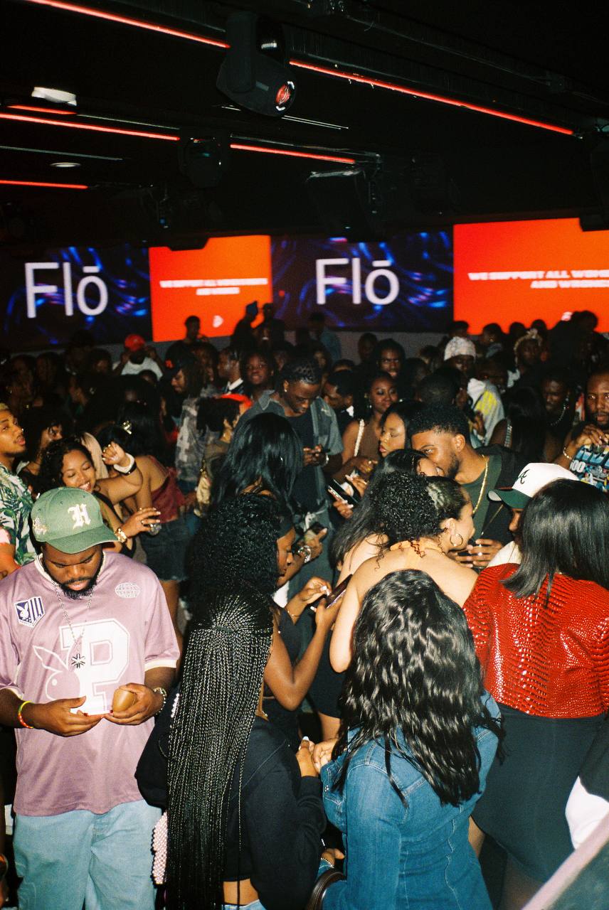 Crowd of people at an indoor event with screens displaying 'Flo' and red graphics, some using phones.