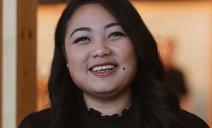 Smiling woman with long dark hair in a dimly lit indoor setting.