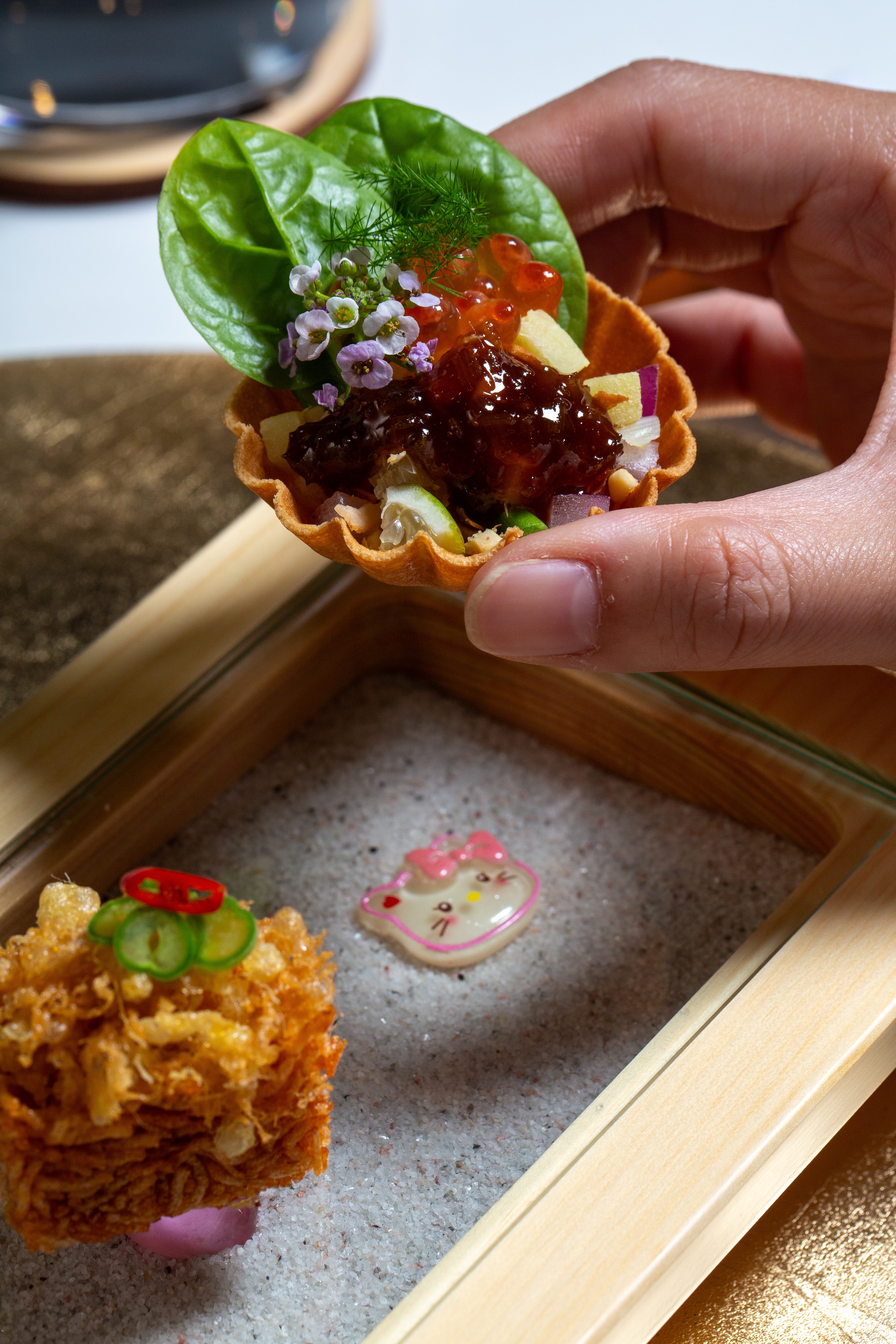 Hand holding a small edible cup with lettuce, red sauce, and toppings, above a wooden box with a decorated cookie and fried food.