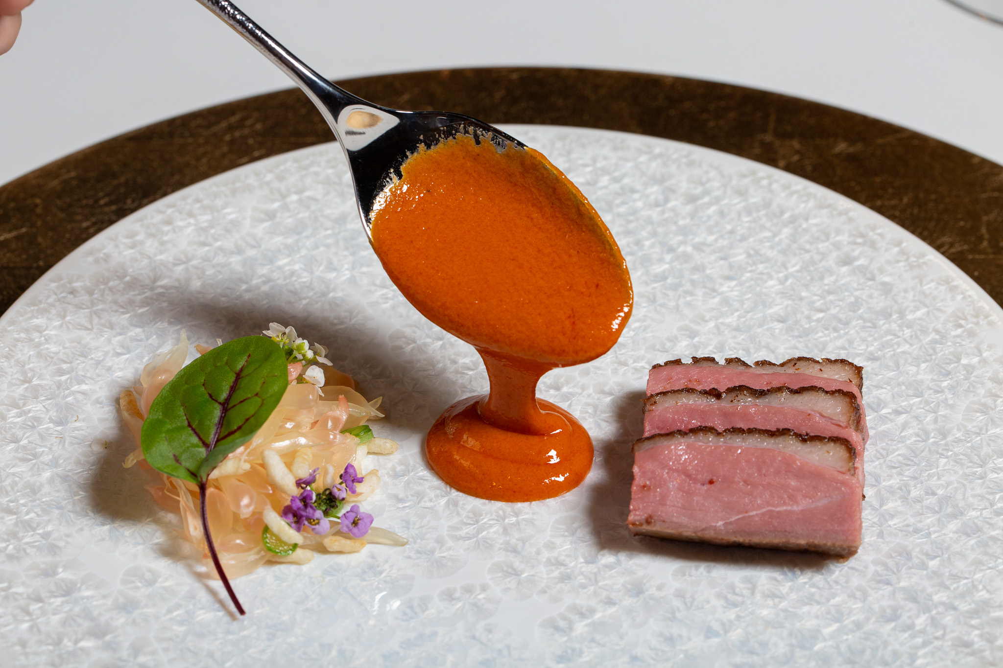 Spoon with orange sauce, a small green leaf, and a pink meat dish on a white plate.